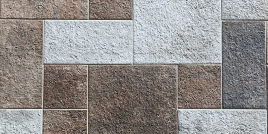 A close-up of a textured, multi-colored stone tile pattern. Rectangular and square tiles in various shades of gray, brown, and beige are arranged in a staggered, interlocking layout. The rough substrate of each tile features a grainy texture.